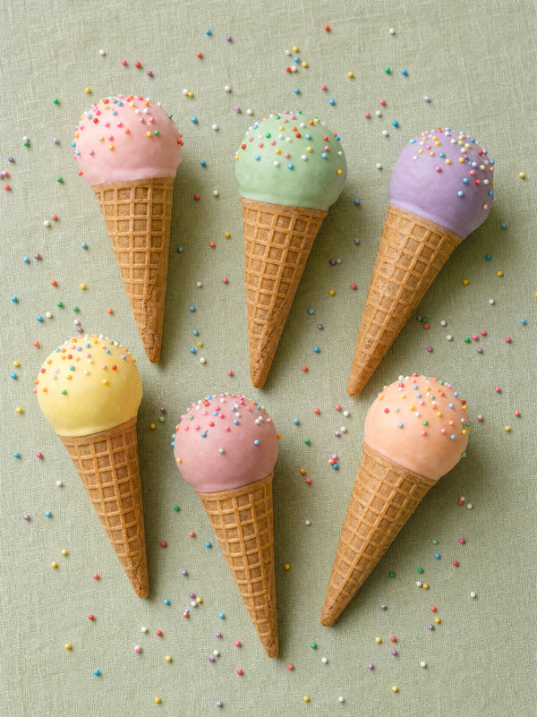 Pastel cake pops in mini waffle cones on a sage surface, sprinkled with rainbow nonpareils